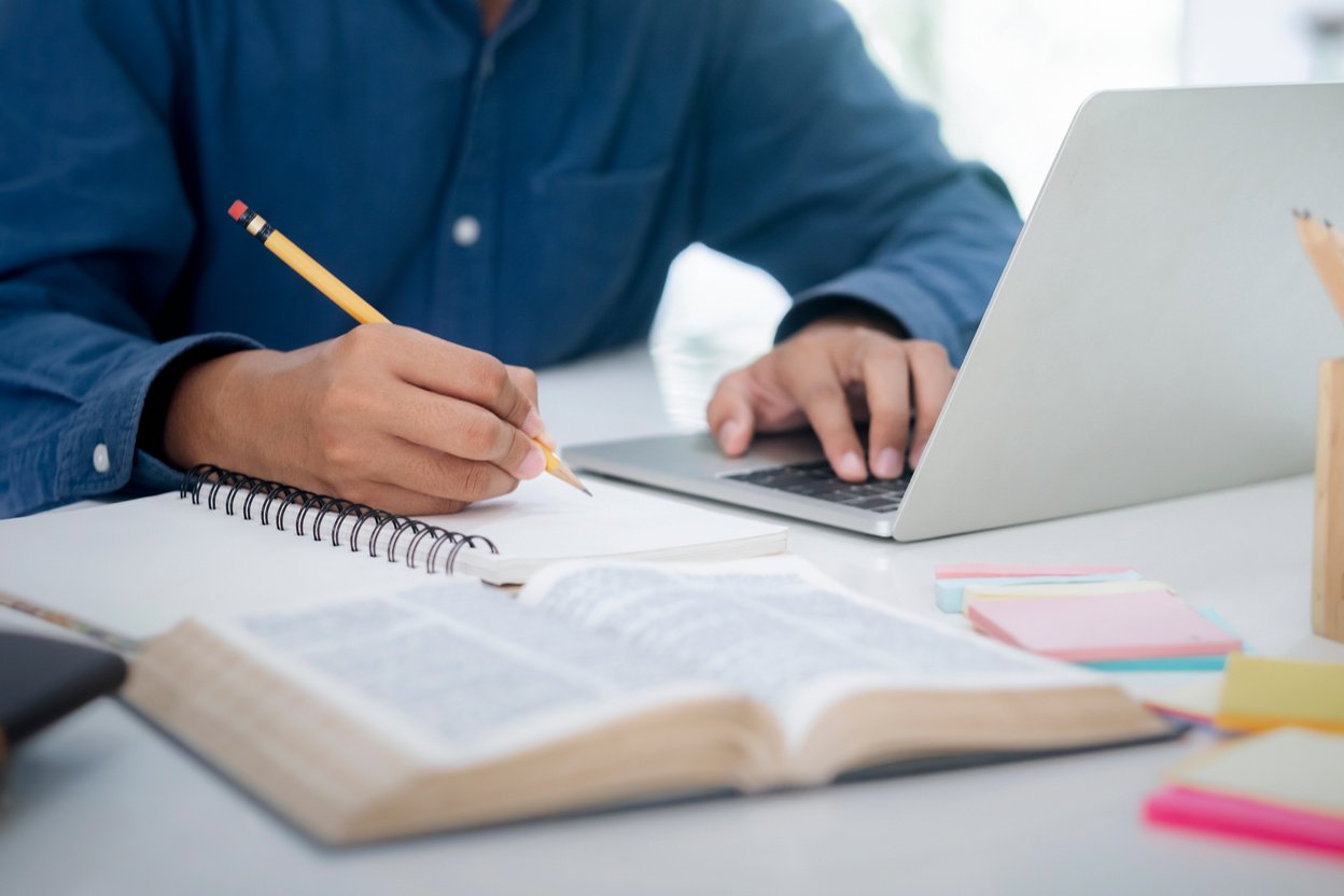 Male Student Writing on His Notebook while Doing Online Research